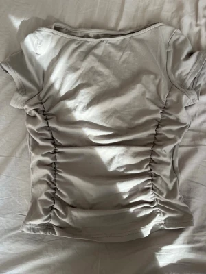 Grå ribbad topp med rynkade sidor - Snygg grå topp med korta ärmar och rynkade sidor som ger en cool effekt. Toppen har en rund halsringning och är gjord i mjuk bomull. Perfekt att styla med jeans eller kjol för en trendig look.