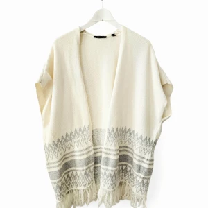Off-white poncho från Esprit - Off-white stickad poncho med öppen framsida från ESPRIT. Den har en lös, ärmlös design med grå geometriska mönster nära nederkanten och fransar längs fållen. Beskrivning av materialet i artikeln som visas på bilden.