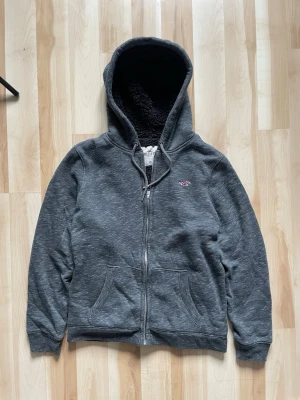 Grå zip hoodie från Hollister - Mysig grå hoodie från Hollister med dragkedja och två fickor framtill. Insidan är fodrad med svart teddyfoder för extra värme. Klassisk huva med dragsko och liten Hollister-logga broderad på bröstet. Perfekt för chill dagar.