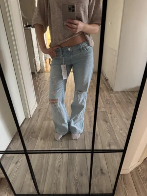 Ljusblå ripped jeans med raka ben - Säljer ett par ljusblå jeans med raka ben och slitna detaljer på låren och knäna. Jeansen har hög midja och en avslappnad passform. Perfekta för en chill och trendig look. Materialet är bomull och de har klassisk femficksdesign.