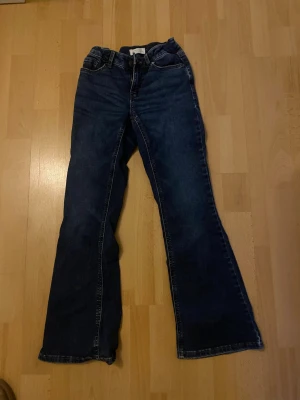 Mörkblå bootcut jeans från Lindex - Säljer ett par mörkblå bootcut jeans från Lindex med klassisk femficksdesign och kontrastsömmar. Jeansen har normal midja och är tillverkade i denim med lätt stretch. Perfekta för en avslappnad och trendig look. Storlek 146