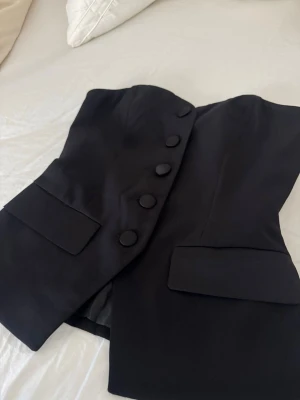 Svart korsettopp med knappar - Snygg svart korsettopp med stora klädda knappar framtill och två dekorativa fickor. Toppen är axelbandslös och har en figurnära passform som ger en elegant siluett. Perfekt att styla med jeans eller kjol för en trendig look.