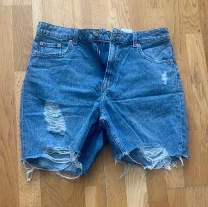 Blå jeansshorts med slitningar - Säljer ett par blå jeansshorts med råa, fransiga benslut och slitna detaljer framtill. Klassisk femficksmodell med hög midja och dragkedja. Perfekta för en avslappnad och trendig look.