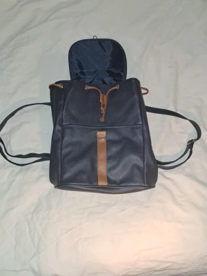 Ryggsäck med bruna detaljer - Marinblå ryggsäck/väska i stilren design med bruna detaljer. Väskan har justerbara axelband och stängs med snörning samt lock. Perfekt för vardag, skola eller mindre utflykter. (Den Väger 40g)