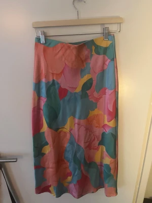 Färgglad blommig långkjol - Säljer en långkjol med stort blommigt mönster i rosa, orange, grön, gul och blå. Kjolen har en rak passform och är tillverkad i ett glansigt, mjukt material som ger en lyxig känsla. Perfekt för dig som vill sticka ut med färg och mönster.