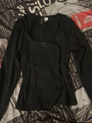 Svart ribbad topp med rosett - Säljer en svart långärmad topp. Toppen är ribbad med en liten söt rosett framtill vid urringningen och har en figurnära passform. Perfekt att styla med jeans eller kjol för en enkel men snygg look.