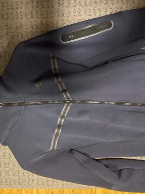 Mörkblå Nike tech fleece - Snygg mörkblå hoodie med hel dragkedja framtill och huva. Hoodien har diskreta reflexdetaljer över bröstet och en liten logga på vänster sida. Perfekt för dig som gillar en stilren och sportig look. Materialet känns mjukt och bekvämt. Pris kan diskuteras