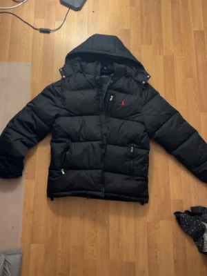 Svart dunjacka från Polo Ralph Lauren - Snygg svart dunjacka från Polo Ralph Lauren med huva och dragkedja. Jackan har två framfickor med dragkedja och en bröstficka, samt det klassiska röda Polo-logotypen broderad på bröstet. Perfekt för kalla vinterdagar.