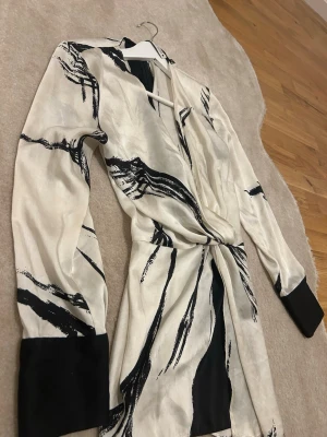 Beige omlottblus med svart mönster - Trendig beige omlottblus med svart abstrakt mönster och breda svarta muddar vid ärmsluten. Blusen har långa ärmar och knytdetalj i midjan som ger en snygg siluett. Materialet ser ut att vara mjukt och glansigt, perfekt för en cool och modern look.