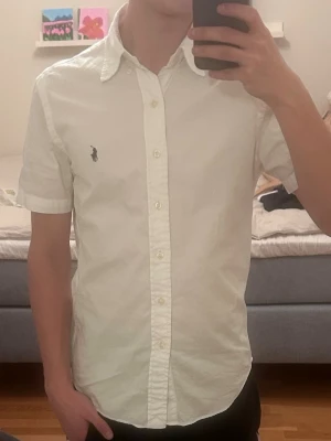 Vit kortärmad skjorta från Ralph Lauren - Vit kortärmad skjorta från Ralph Lauren med klassisk krage och knappar framtill. Skjortan har en broderad logotyp på bröstet och är tillverkad i bomull. Såklart äkta, priset är ej hugget i sten, hör gärna av vid intresse. MVH Erik