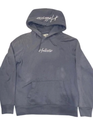 Mörkgrå hoodie från Hollister - Snygg mörkgrå hoodie från Hollister med vit broderad logga på bröstet och huva med dragsko. Klassisk känguruficka framtill och ribbade muddar vid ärmslut och nederkant. Perfekt för en avslappnad och trendig stil.