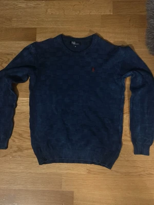 Mörkblå stickad tröja från Polo Ralph Lauren - Säljer en mörkblå stickad tröja från Polo Ralph Lauren med diskret rutmönster och röd broderad logga på bröstet. Tröjan har rund halsringning och ribbade muddar vid ärmslut och nederkant. Perfekt för dig som gillar klassisk och stilren design.