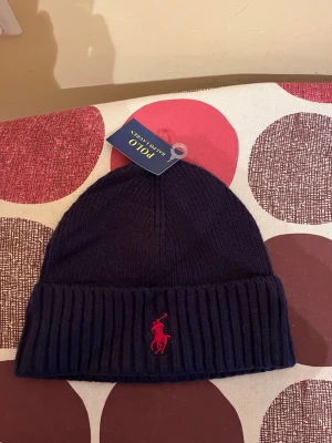 Mörkblå mössa från Polo Ralph Lauren - Snygg mörkblå ribbstickad mössa från Polo Ralph Lauren med uppvikt kant och klassiska röda logotypen broderad framtill. Perfekt accessoar för kalla dagar och enkel att matcha med olika outfits. Tillverkad i mjukt material för extra komfort.