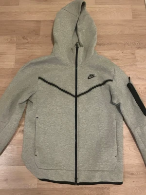 Grå Nike Tech Fleece hoodie - Snygg grå Nike Tech Fleece hoodie med hel dragkedja och svarta detaljer. Har en huva, fickor framtill och en extra ficka med dragkedja på ärmen. Tillverkad i mjukt och bekvämt material, perfekt för en sportig och avslappnad stil. Helt ny knappast använd. Storlek S men passar för M också.