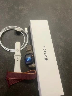 Apple Watch med extra armband - Apple Watch med svart fyrkantig urtavla, vit silikonrem, extra sportloop i vinrött och orange samt ett metallnätarmband i guld. Laddkabel och originalförpackning ingår. Perfekt för dig som vill ha koll på träning, notiser och hälsa direkt på handleden.