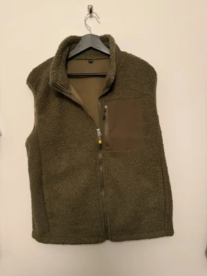 Olivgrön teddyväst med dragkedja - Mysig olivgrön teddyväst med hög krage och hel dragkedja framtill. Västen har en praktisk bröstficka i matchande tyg med dragkedja. Perfekt för lager-på-lager och ger en cool, avslappnad look.