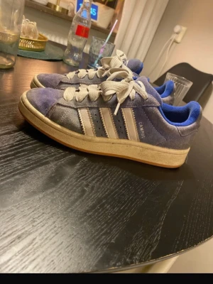 Adidas Campus blå sneakers - Säljer ett par klassiska Adidas Campus sneakers i blå mocka med vita detaljer och breda vita snören. Skorna har de ikoniska tre ränderna på sidan och Adidas-logga på plösen. Låg modell med vit sula och bekväm passform. Perfekta för en avslappnad streetstyle. Skick se bilder😊. Solblekta. Hör av er vid frågor💞😊