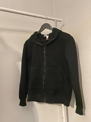 Svart hoodie från C.P. Company - Snygg svart hoodie från C.P. Company med dragkedja framtill och broderad logga på bröstet. Hoodien har två stora fickor och en klassisk huva. Tillverkad i mjukt material som känns skönt mot huden. Perfekt för en avslappnad och stilren look.