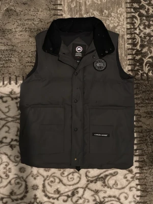 Svart dunväst från Canada Goose - Snygg svart dunväst från Canada Goose med hög krage och tryckknappar framtill. Västen har två stora framfickor och en diskret logotyp på bröstet samt en patch med märkesnamnet på fickan. Perfekt för lager-på-lager och streetwear.