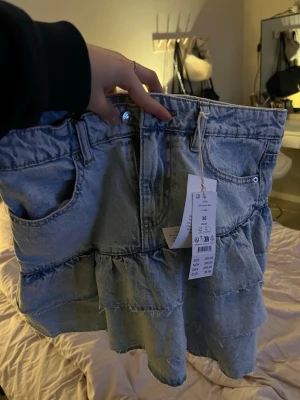 Volangkjol i ljus denim från Gina - Säljer en kort volangkjol i ljusblå denim från Gina Tricot. Kjolen har klassiska jeansdetaljer med fickor fram och bak, knapp och dragkedja samt flera lager volanger💗