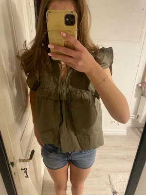 Olivgrön blus med volangdetaljer - Säljer en olivgrön blus med korta ärmar och v-ringning. Blusen har volangdetaljer och en lös passform som ger en avslappnad look. Perfekt att matcha med jeansshorts eller kjol för en trendig stil.