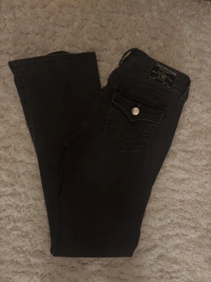 Gråa True Religion bootcut jeans - Säljer ett par Gråa True Religion BECCA Mid Rise Bootcut jeans med snygga kontrastsömmar och ikonisk fickdetalj med knapp. Jeansen har medelhög midja och bootcut passform, perfekta för en avslappnad men ändå trendig look.
