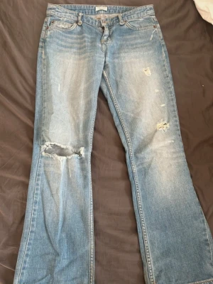Blå bootcut jeans med slitningar - Snygga blå bootcut jeans med slitna detaljer och hål på benen. Jeansen har klassiska bakfickor med dekorativa sömmar och en ljus tvätt. Perfekta för en avslappnad och trendig look.