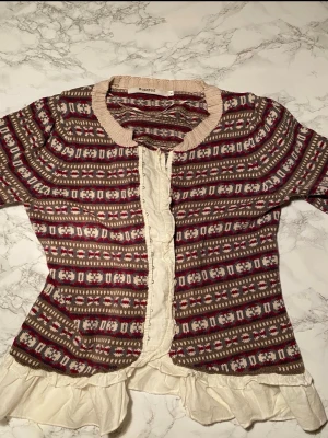 Mönstrad bomullskofta från Rosebud - Unik kofta från Rosebud 100% bomull  med beige, vinrött mönster. Tröjan har ribbstickade muddar och krage i beige samt dekorativa volanger i vitt tyg längs knappslån och nederkanten. Perfekt för dig som gillar vintageinspirerad stil.