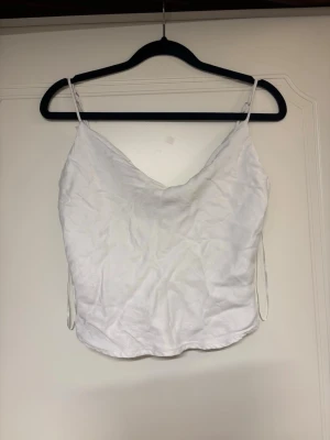 Vit satin topp med knappar - Vit satin topp med tunna axelband och lätt glansigt material. Toppen har en snygg draperad v-ringning och dekorativa knappar baktill. 