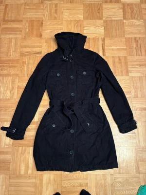 Svart trenchcoat med bälte - Stilren svart trenchcoat med hög krage, knäppning framtill och bälte i midjan. Jackan har två bröstfickor med lock, två snedställda fickor och justerbara spännen vid ärmsluten. Perfekt för en cool och klassisk look.