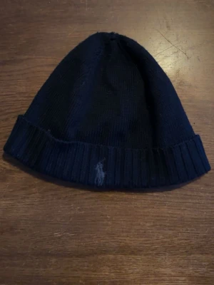 Mörkblå mössa från Polo Ralph Lauren - Snygg mörkblå ribbstickad mössa från Polo Ralph Lauren med den klassiska broderade loggan framtill. Tillverkad i mjukt material som håller dig varm och passar perfekt till kalla dagar. Enkel och stilren design som funkar till det mesta.