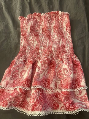 Röd och vit bandeau tubtopp med volang - Supersöt bandeau tubtopp i rosa och vitt med paisleyinspirerat mönster och volangdetaljer. Toppen är ärmlös och har elastisk smock upptill för skön passform. Perfekt för sommarens varma dagar och festivaler.