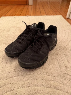 Nike Air Max Plus Tn  - Säljer ett par helsvarta Nike Air Max Plus Tn sneakers med klassisk mesh- och syntetöverdel, svarta snören och ikonisk Tn-detalj på hälen. Skorna har en chunky sula med synlig Air-enhet och vågiga detaljer på ovandelen. Perfekta för dig som gillar streetwear.