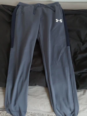 Grå mjukisbyxor från Under Armour - Snygga grå mjukisbyxor från Under Armour med mörkblå sidopaneler och dragsko i midjan. Byxorna har resår vid bensluten och ett vitt Under Armour-logo tryck på ena benet. Perfekta för chill eller träning. Storlek 160-170 cm 