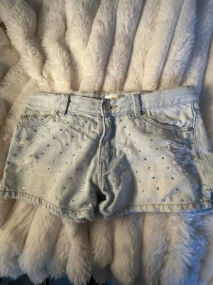 Ljusblå denimshorts med nitar - Snygga ljusblå dhorts från ginatricot  med silverfärgade pärlor framtill och slitna detaljer. med bälteshällor och knappgylf. Perfekta för varma dagar och ger en cool vibe till din outfit. de går att ändra i midian och har ingenting på bakfickorna💗