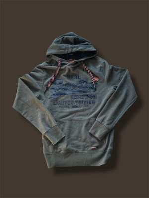 Olivgrön hoodie från Superdry - Snygg olivgrön hoodie från Superdry med stor logga och text på bröstet. Hoodien har huva med röda och vita snören samt en klassisk känguruficka framtill. Tillverkad i mjuk bomullsblandning och har ribbade muddar vid ärmslut och nederkant. Det står storlek M men den är ganska liten i  storlek så passar bättre som storlek S. Modellen är 180 och väger 65