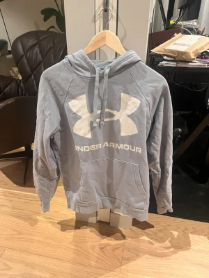 Ljusblå hoodie från Under Armour - Snygg ljusblå hoodie från Under Armour med stor vit logga och text på bröstet. Hoodien har huva med dragsko och en stor magficka framtill. Tillverkad i mjukt material som känns skönt mot huden. Perfekt för en avslappnad och sportig stil.