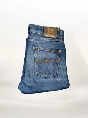 Blå jeans från Nudie Jeans - Säljer ett par klassiska blå jeans från Nudie Jeans med snygga slitningar och kontrastsömmar. Jeansen har fem fickor, rak passform och är gjorda i slitstarkt denim. Perfekta för dig som gillar en tidlös och avslappnad stil.