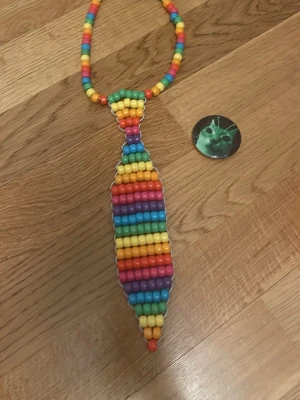 Färgglad pärlfluga till maskerad - Unik slips till maskerad gjord av plastpärlor i regnbågens färger. Slipsen har ett halsband av pärlor och själva slipsen är vävd i ränder av gult, orange, rött, blått, grönt och lila. Perfekt accessoar för cosplay eller temafest.