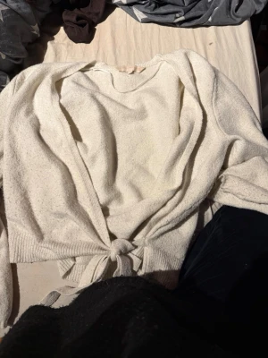 Krämvit omlottkofta med knytband - Mysig krämvit omlottkofta med knytband i midjan och långa ärmar. Koftan har en avslappnad passform och ribbade muddar vid ärmslut och nederkant. Perfekt att slänga över en topp för en chill look.