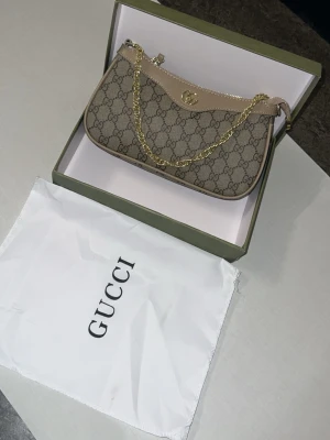 Gucci beige monogram handväska - Säljer en snygg beige handväska från Gucci med klassiskt GG-monogram i canvas och detaljer i ljusbrunt läder. Väskan har guldfärgad kedja och justerbar axelrem. Perfekt accessoar för dig som gillar lyxig och trendig stil.