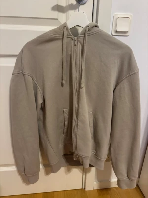 Beige zip hoodie med huva - En stilren beige hoodie med dragkedja framtill och huva med dragsko. Hoodien har två fickor på magen och ribbade muddar vid ärmar och nederkant. Perfekt för en avslappnad och enkel look.