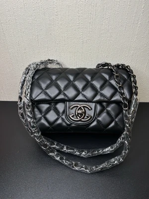 Svart Chanel axelväska i läder - Snygg svart axelväska från Chanel i quiltat läder med silverfärgat CC-spänne framtill. Kedjerem med insytt läder ger en lyxig känsla. Klassisk och tidlös design som passar till många olika stilar.