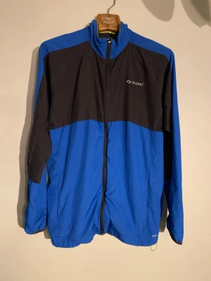 Blå och svart vindjacka från SOC - Sportig vindjacka från SOC i blått och svart med dragkedja framtill. Jackan har hög krage, elastiska muddar och reflekterande detalj på ryggen. Tillverkad i lätt och vindtätt material, perfekt för utomhusaktiviteter. Logga på bröstet.