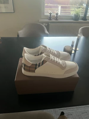 Burberry skor - Snygga vita sneakers från Burberry med klassiskt beige, svart och rött rutmönster på hälen. Skorna har vita snören och detaljer i beige mocka. Perfekta för dig som vill ha en clean och stilren look med en touch av lyx.
