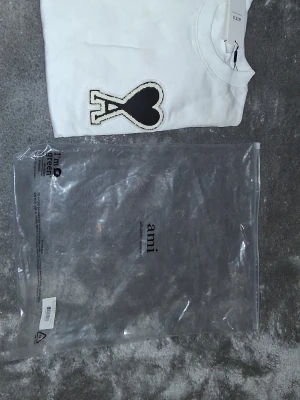 Vit t-shirt från Ami med hjärtdetalj - Snygg vit t-shirt från Ami med stor svart hjärtdetalj på bröstet. Tillverkad i mjuk bomull och har en klassisk rund halsringning. Perfekt för dig som gillar stilrena plagg med en twist.