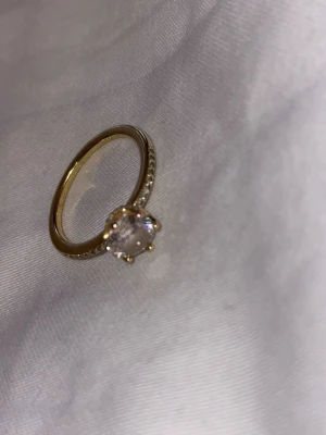 Guldfärgad Promise ring med stor sten - Snygg guldfärgad ring med en stor, rund klar sten i mitten och små gnistrande stenar längs bandet. Perfekt för dig som gillar bling och vill sticka ut med dina accessoarer. Klassisk och elegant design som passar till många stilar. St 54 värde 1199kr 