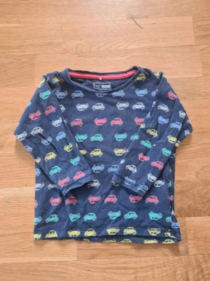 Blå långärmad tröja med bilar - Mysig långärmad barntröja från name it i storlek 80 i mörkblått med färgglada bilmotiv över hela plagget. Tröjan har rund halsringning och är perfekt för dig som gillar lekfulla mönster. Materialet känns mjukt och bekvämt mot huden.
