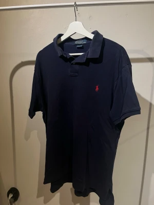 Mörkblå pikétröja från Ralph Lauren - Klassisk mörkblå pikétröja från Ralph Lauren med röd broderad logga på bröstet. Tröjan har krage, korta ärmar och knäppning vid halsen. Perfekt för en stilren och avslappnad look.