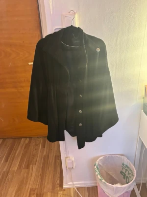 Svart cape-kappa från Stockh LM - Snygg svart cape-kappa från Stockh LM med knappar framtill och bred krage. Kappan har en lös och flödande passform som ger en cool siluett. Perfekt för dig som vill ha en stilren och trendig look. Materialet känns mjukt och följsamt.